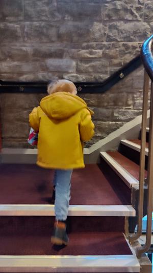 A small boy in a yellow jacket climbs a set of red carpeted stairs 