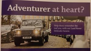 Photo of a print advert. A land rover is driving on a cobbled street. The caption reads "Help them remember the real you, with our Land Rover Defender hearse" 